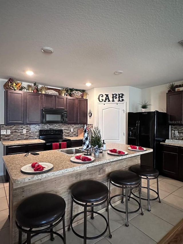 kitchen with an island with sink, a kitchen breakfast bar, light tile patterned floors, black appliances, and dark brown cabinets