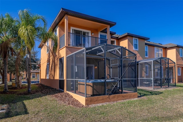 back of house with glass enclosure, a hot tub, a balcony, and a yard