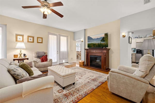 living area with visible vents, a ceiling fan, a glass covered fireplace, light wood-type flooring, and baseboards