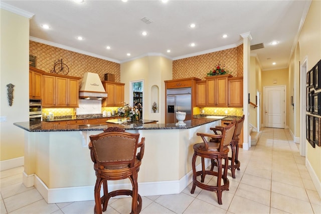 kitchen with built in fridge, dark stone countertops, a breakfast bar area, a large island with sink, and premium range hood