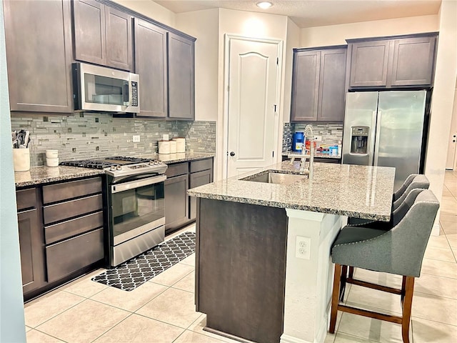 kitchen with a kitchen bar, backsplash, a kitchen island with sink, appliances with stainless steel finishes, and light stone counters