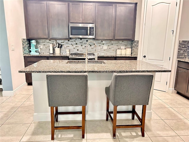 kitchen with a kitchen bar, a kitchen island with sink, light tile patterned floors, light stone counters, and dark brown cabinetry