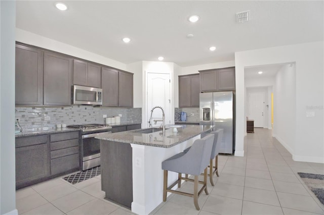 kitchen with light tile patterned floors, sink, dark stone counters, stainless steel appliances, and an island with sink