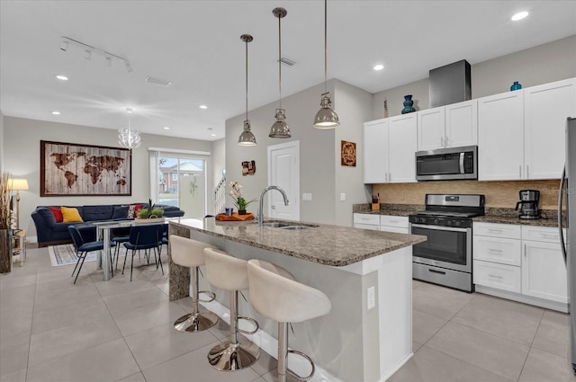 kitchen with an island with sink, stainless steel appliances, dark stone counters, hanging light fixtures, and sink