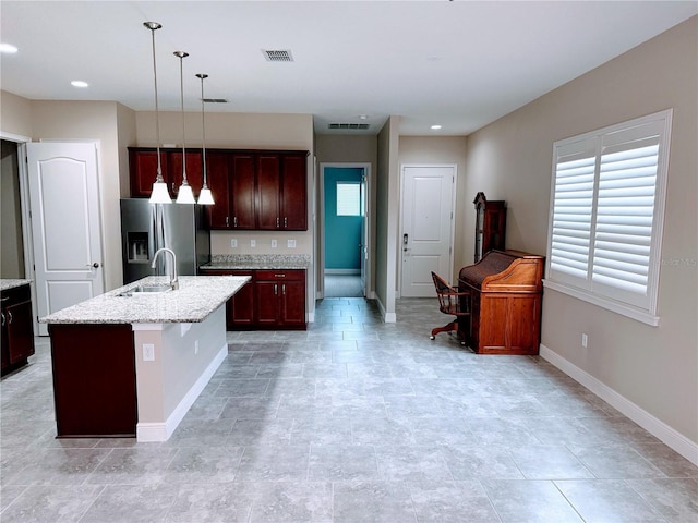 kitchen with an island with sink, stainless steel refrigerator with ice dispenser, pendant lighting, light stone counters, and sink