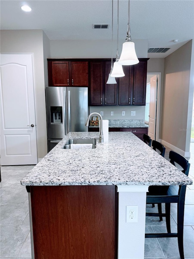 kitchen with light stone countertops, pendant lighting, an island with sink, sink, and stainless steel fridge with ice dispenser