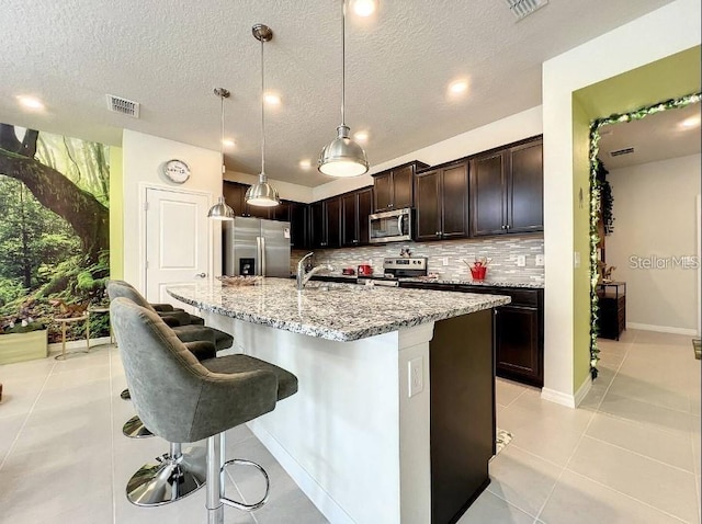 kitchen with decorative light fixtures, stainless steel appliances, an island with sink, light tile patterned floors, and decorative backsplash