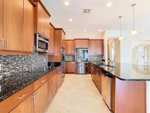 kitchen with pendant lighting, appliances with stainless steel finishes, dark stone counters, sink, and a kitchen island with sink