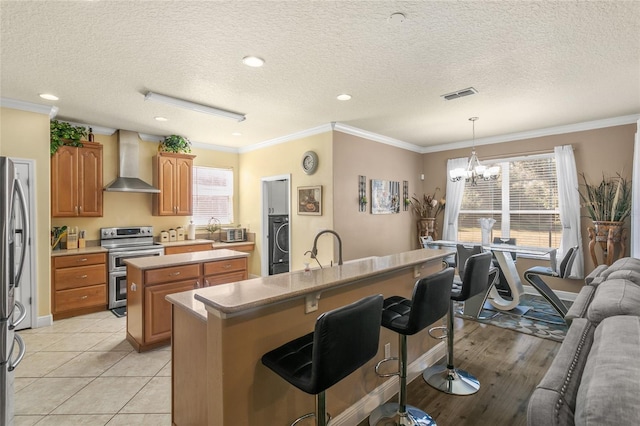 kitchen featuring an island with sink, washer / dryer, stainless steel appliances, a textured ceiling, and wall chimney exhaust hood