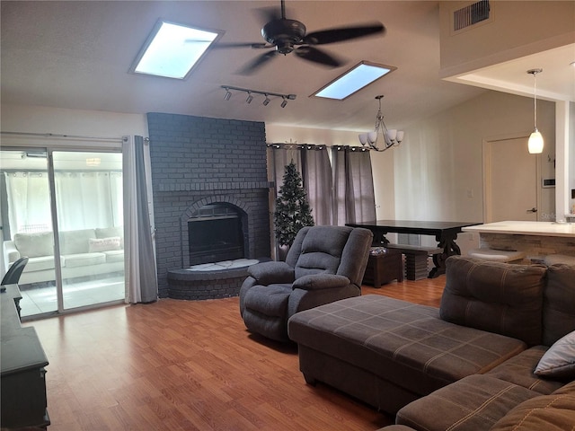 living room with ceiling fan with notable chandelier, vaulted ceiling with skylight, hardwood / wood-style floors, and a brick fireplace