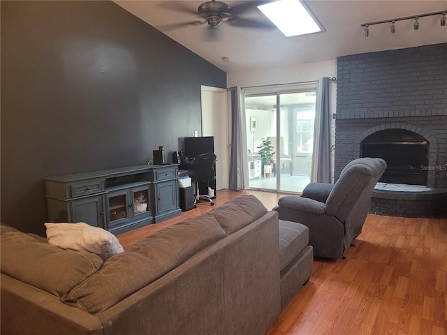 living room with vaulted ceiling, rail lighting, ceiling fan, light hardwood / wood-style floors, and a brick fireplace