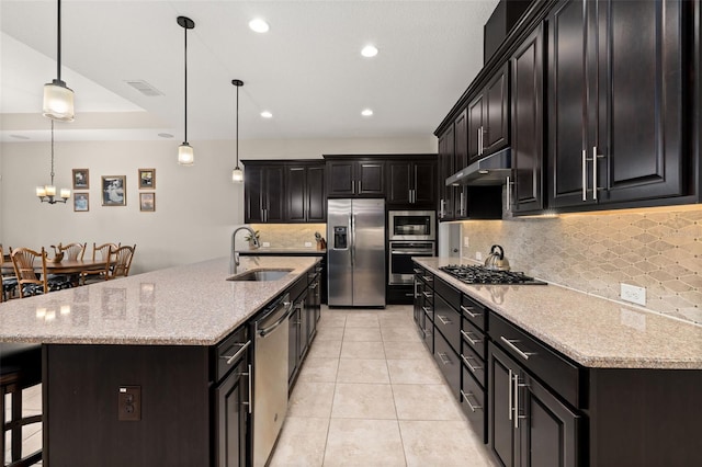 kitchen with appliances with stainless steel finishes, hanging light fixtures, an island with sink, light stone counters, and sink