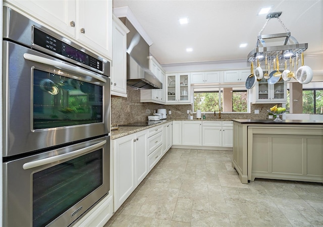 kitchen featuring light stone countertops, stainless steel double oven, white cabinets, wall chimney range hood, and sink