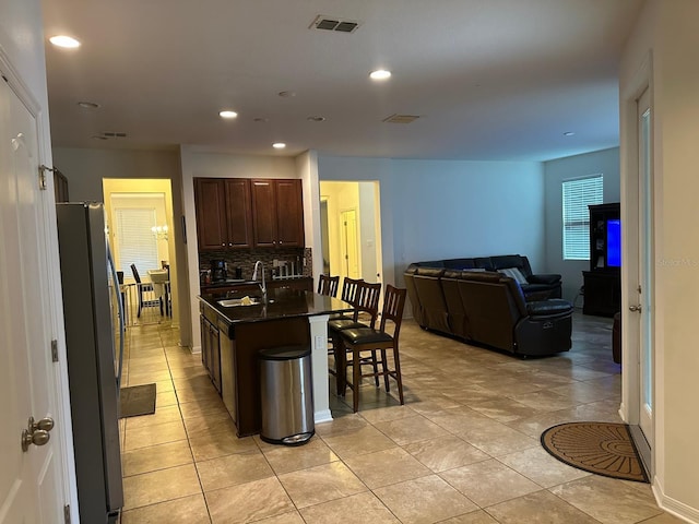 kitchen with sink, light tile patterned floors, a center island with sink, a breakfast bar area, and stainless steel refrigerator