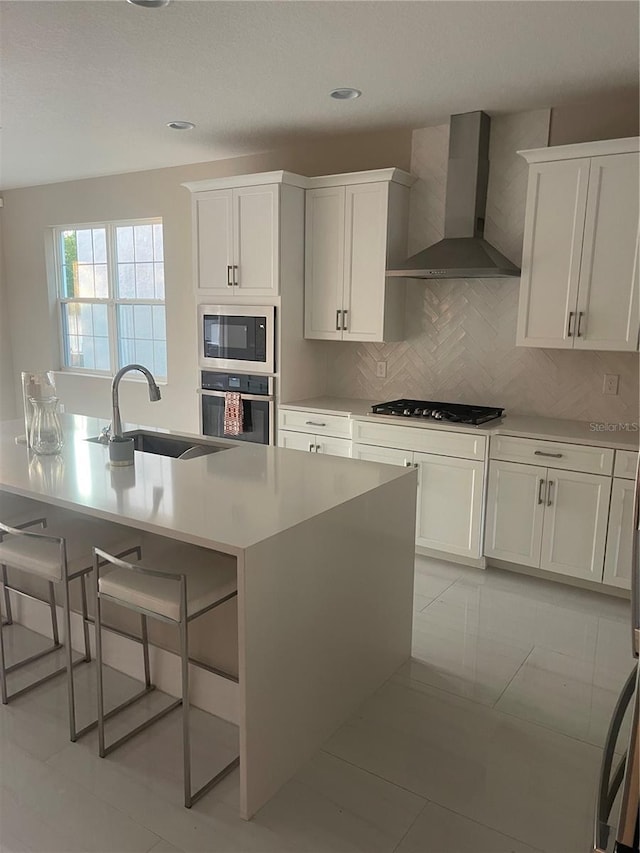 kitchen featuring appliances with stainless steel finishes, wall chimney range hood, sink, backsplash, and a kitchen island with sink