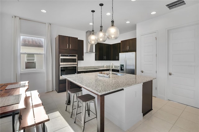 kitchen featuring a center island with sink, stainless steel appliances, hanging light fixtures, light tile patterned floors, and dark brown cabinets