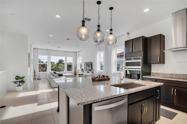 kitchen with appliances with stainless steel finishes, an island with sink, light stone countertops, wall chimney range hood, and light tile patterned flooring