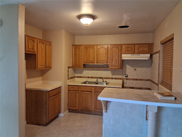 kitchen with light tile patterned flooring, a sink, light countertops, under cabinet range hood, and brown cabinets