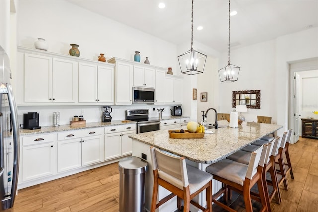 kitchen featuring white cabinets, decorative light fixtures, an island with sink, stainless steel appliances, and light stone counters