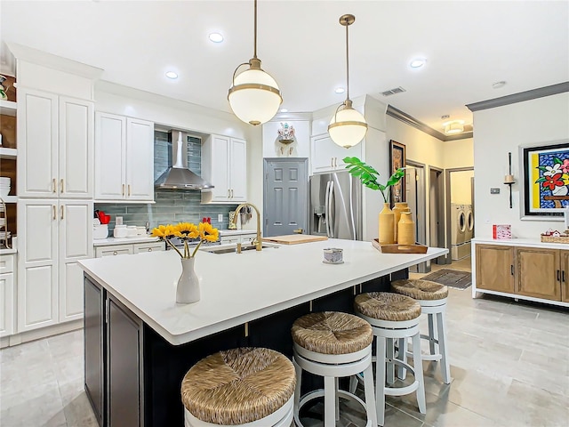 kitchen with stainless steel refrigerator with ice dispenser, white cabinetry, decorative light fixtures, a kitchen island with sink, and wall chimney range hood