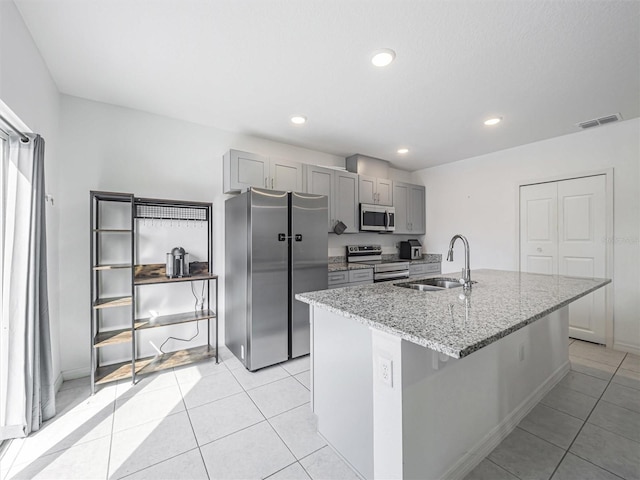 kitchen with gray cabinets, stainless steel appliances, a kitchen island with sink, light stone countertops, and sink