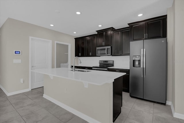 kitchen with sink, light tile patterned flooring, dark brown cabinetry, a kitchen island with sink, and appliances with stainless steel finishes