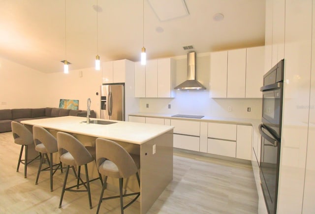 kitchen featuring decorative light fixtures, wall chimney range hood, white cabinets, and a center island with sink