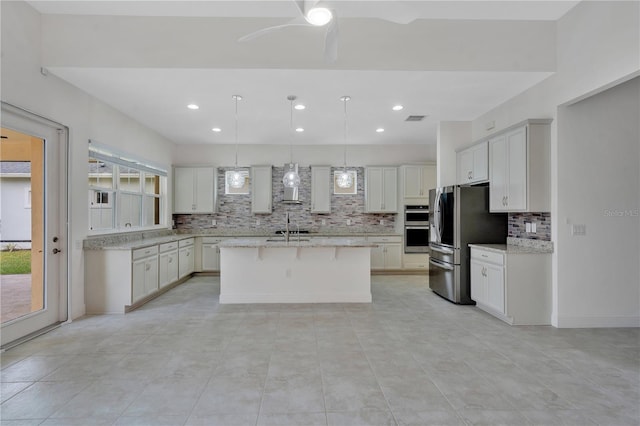 kitchen featuring appliances with stainless steel finishes, wall chimney range hood, hanging light fixtures, ceiling fan, and a center island with sink