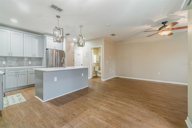 kitchen with tasteful backsplash, decorative light fixtures, light hardwood / wood-style flooring, stainless steel fridge, and an island with sink