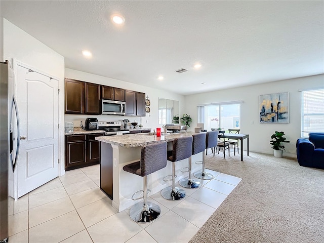 kitchen featuring dark brown cabinetry, a kitchen bar, stainless steel appliances, an island with sink, and light colored carpet