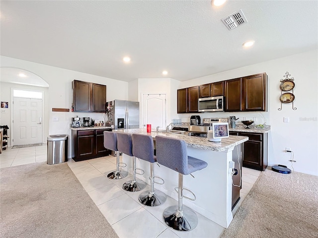 kitchen featuring a kitchen breakfast bar, stainless steel appliances, light tile patterned floors, a center island with sink, and dark brown cabinetry