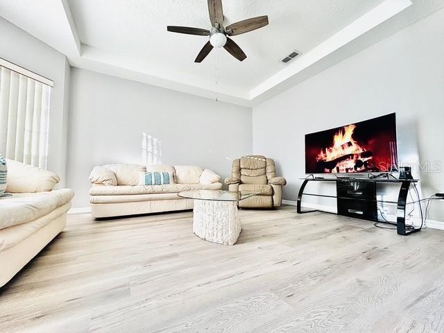 living room with ceiling fan, visible vents, baseboards, a tray ceiling, and light wood finished floors