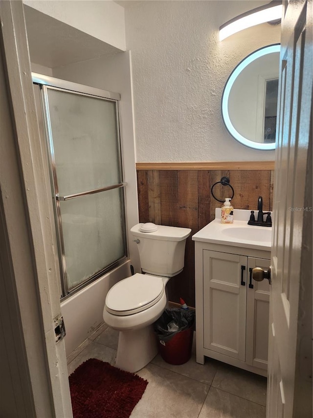 full bathroom with tile patterned flooring, bath / shower combo with glass door, vanity, toilet, and wood walls