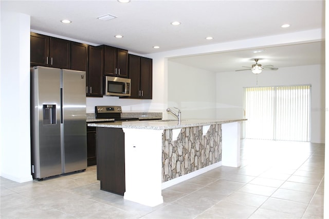 kitchen with light tile patterned floors, ceiling fan, appliances with stainless steel finishes, a kitchen island with sink, and dark brown cabinetry