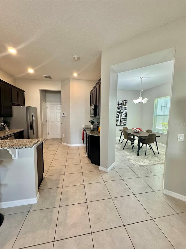 kitchen featuring appliances with stainless steel finishes, light tile patterned floors, dark brown cabinetry, light stone counters, and an inviting chandelier