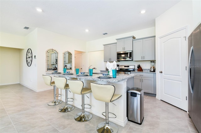 kitchen featuring appliances with stainless steel finishes, a breakfast bar, gray cabinetry, light stone countertops, and a center island with sink