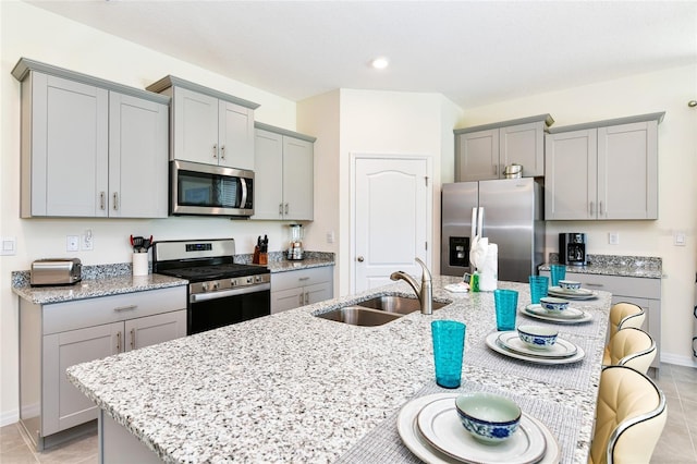 kitchen with sink, a kitchen island with sink, light tile patterned floors, stainless steel appliances, and light stone countertops