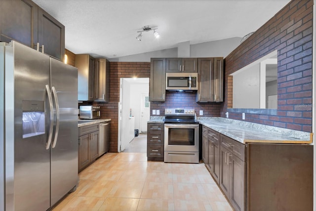 kitchen featuring stainless steel appliances, light stone countertops, brick wall, decorative backsplash, and vaulted ceiling