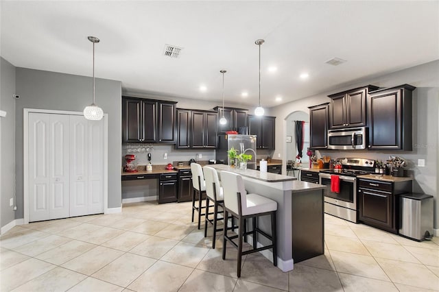 kitchen featuring light tile patterned flooring, dark brown cabinets, appliances with stainless steel finishes, an island with sink, and pendant lighting