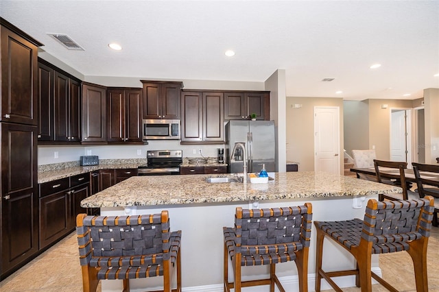 kitchen with appliances with stainless steel finishes, a kitchen island with sink, light stone counters, and dark brown cabinets