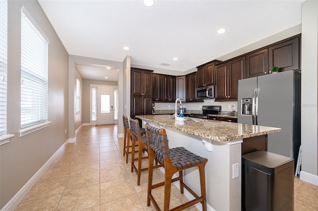 kitchen with appliances with stainless steel finishes, a kitchen bar, a kitchen island with sink, light stone countertops, and dark brown cabinets