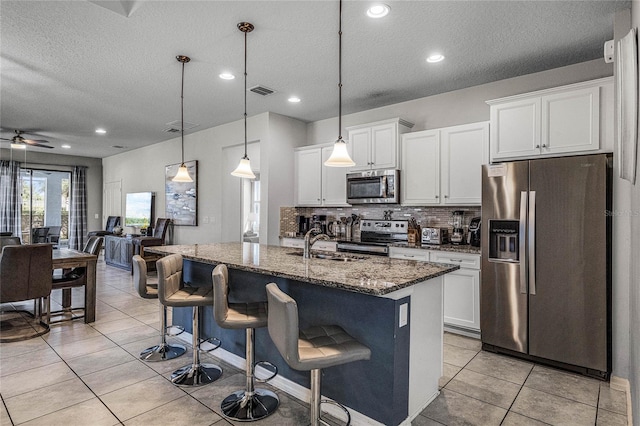 kitchen featuring white cabinets, stainless steel appliances, a center island with sink, and dark stone counters