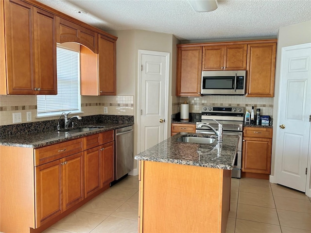 kitchen with sink, appliances with stainless steel finishes, a center island with sink, light tile patterned flooring, and dark stone counters