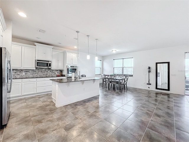 kitchen with dark stone counters, pendant lighting, stainless steel appliances, a kitchen island with sink, and white cabinets