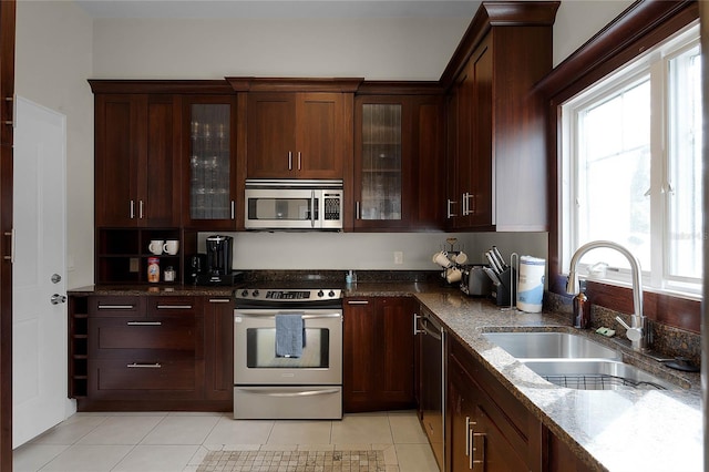 kitchen with light tile patterned floors, a sink, appliances with stainless steel finishes, dark stone counters, and glass insert cabinets