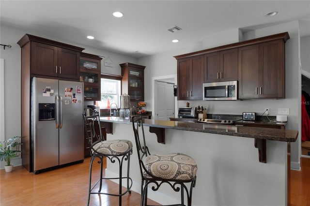 kitchen with visible vents, appliances with stainless steel finishes, glass insert cabinets, dark stone counters, and a kitchen breakfast bar