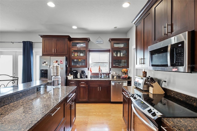 kitchen with stainless steel appliances, a sink, light wood-style floors, dark stone countertops, and glass insert cabinets