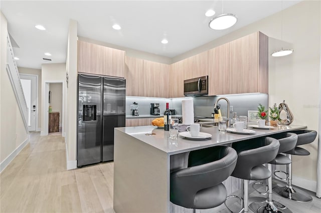 kitchen with appliances with stainless steel finishes, a kitchen bar, hanging light fixtures, light brown cabinets, and light wood-type flooring