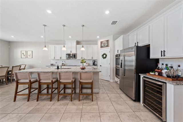 kitchen featuring pendant lighting, stainless steel appliances, wine cooler, white cabinets, and a center island with sink