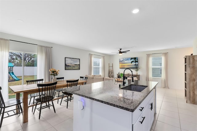 kitchen with an island with sink, sink, dark stone countertops, white cabinets, and light tile patterned floors